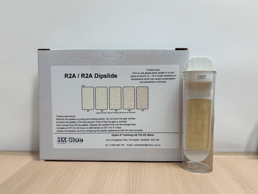 A box of R2A / R2A Dipslide with a clear cylindrical sample vial containing a beige liquid, placed on a light wood surface against a plain white wall.
